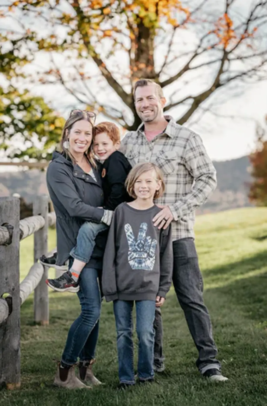 A smiling family of four stands outdoors on grass near a wooden fence. Autumn trees in the background. The mood is joyful and relaxed.