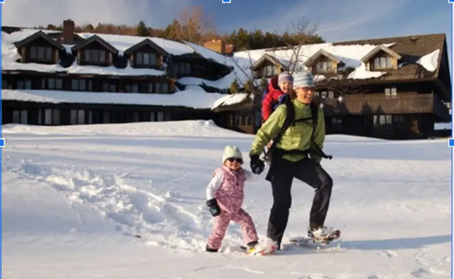 A person snowshoeing in deep snow carries a child in a backpack, another child walks alongside. A large lodge with snowy roofs is in the background.
