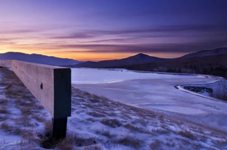 Snow-covered landscape at sunset, with a wooden fence leading to a frozen lake. The sky is a gradient of purple and orange, evoking tranquility and calm.
