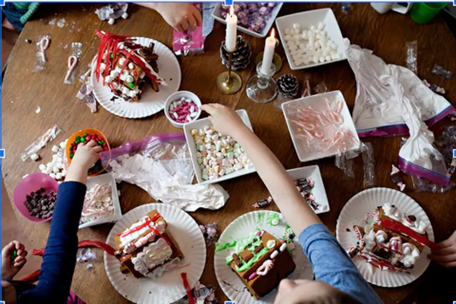 A group of children decorates gingerbread houses with colorful candies and icing on a cluttered wooden table. The scene feels festive and creative.