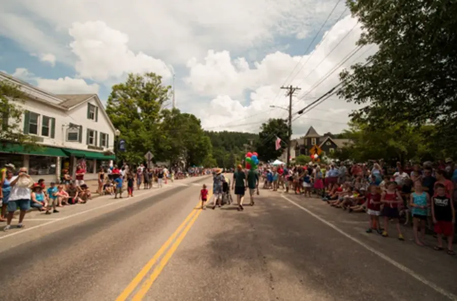 Crowds line a small town street under a partly cloudy sky, anticipating a parade. People hold colorful balloons, creating a festive, cheerful atmosphere.