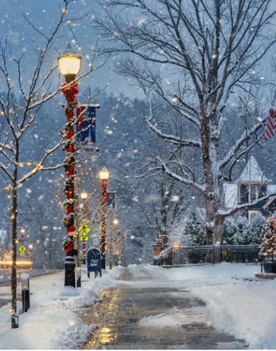 A snow-covered street lined with festive lamp posts wrapped in red garlands. Snow falls gently, creating a peaceful winter scene in a small town.