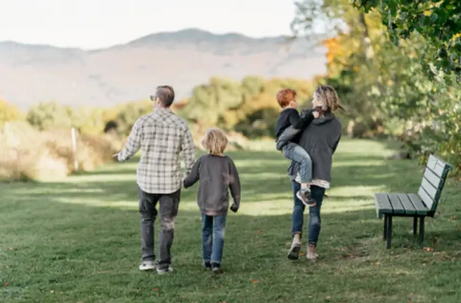 A family of four walks on a grassy path with mountains and autumn trees in the background. One carries a child, evoking a sense of togetherness and tranquility.