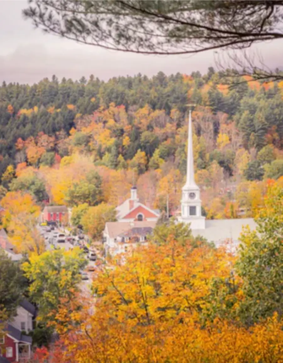 A quaint town is enveloped in vibrant autumn foliage. A white church with a tall spire stands amidst colorful trees, under a cloudy sky, evoking serenity.