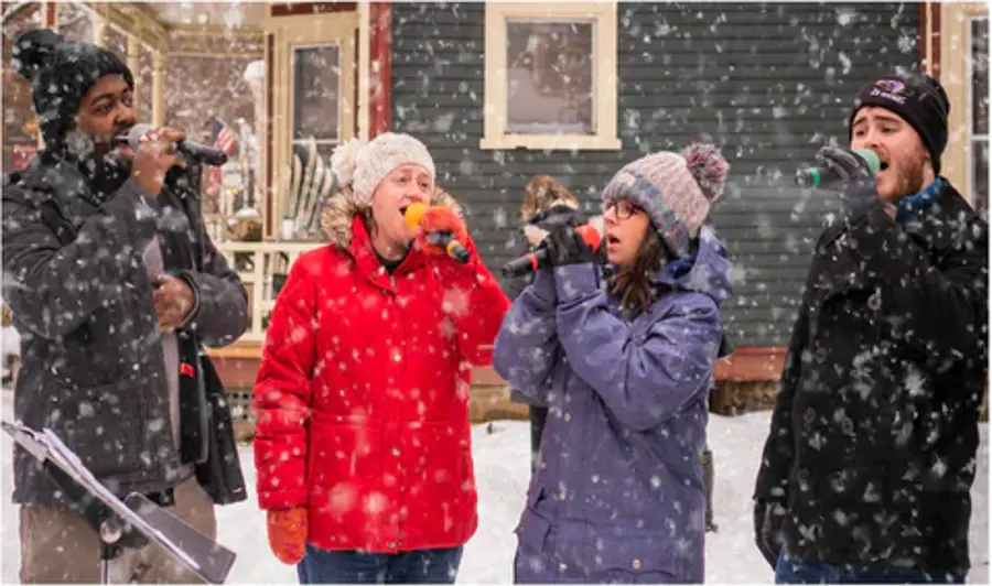 Four people wearing winter clothing sing into microphones outside during a snowfall. They appear joyful and are in front of a dark house in a snowy setting.