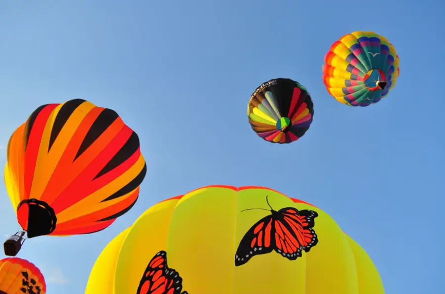Four colorful hot air balloons float in a clear blue sky. One features vibrant stripes, another depicts butterflies on yellow. The scene is lively and cheerful.