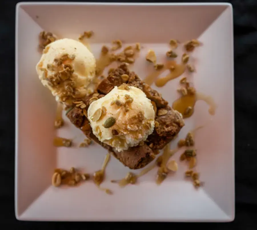 A square plate holds two ice cream scoops on a slice of cake, garnished with nuts and drizzled with caramel, creating a cozy and indulgent vibe.