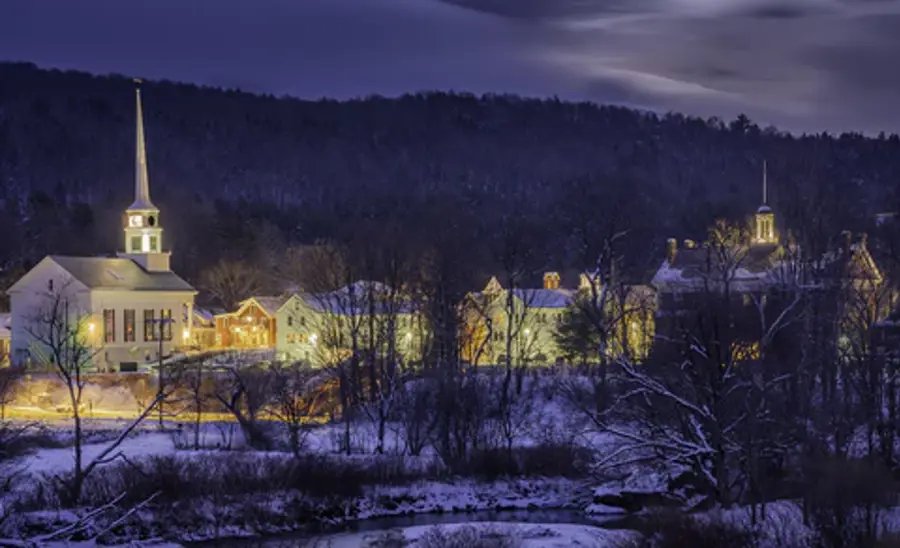 A snowy village at dusk with warmly lit buildings, including a church with a tall steeple. The dark forested hills create a peaceful winter scene.
