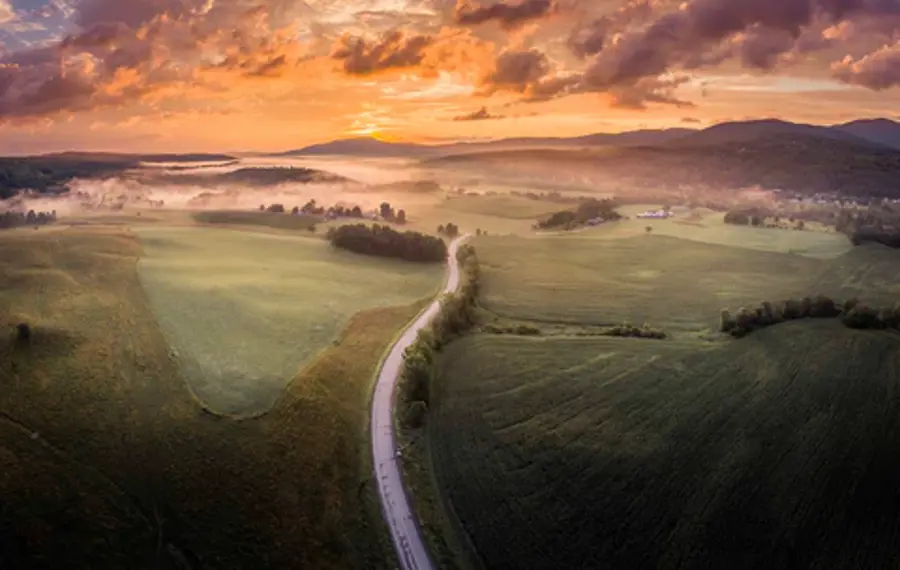Aerial view of a tranquil countryside at sunrise with a winding road through green fields. Mist drifts over distant trees under a sky with warm, glowing clouds.