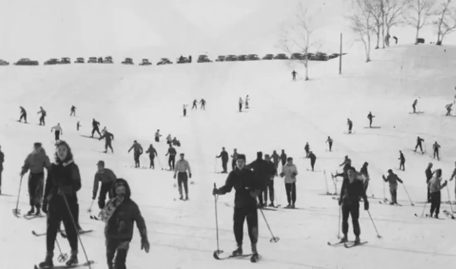 Historic black and white photo of a lively ski slope with numerous skiers, trees in the background, and a line of parked cars atop the hill.
