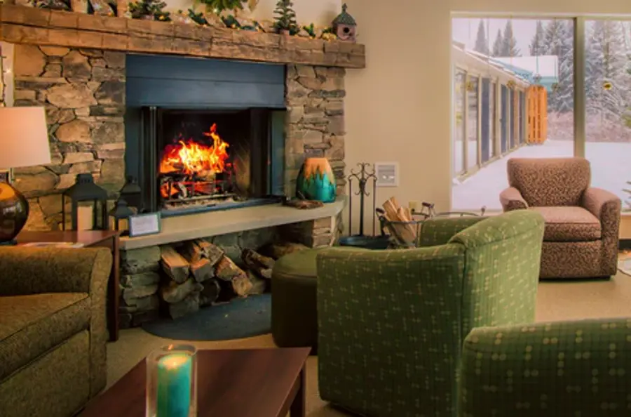 Cozy living room with a roaring stone fireplace, green chairs, and view of snowy landscape through large windows, conveying warmth and comfort.