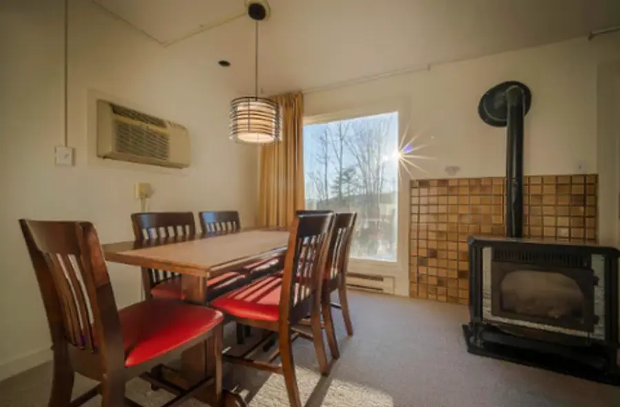 A cozy dining room with a wooden table and six red-cushioned chairs, sunlit window, hanging lamp, wall-mounted AC, and a black wood stove on tiled wall.