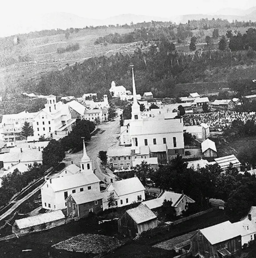 Aerial black-and-white photo of a 19th-century town with prominent church steeples, clustered buildings, and rolling hills in the background. Peaceful atmosphere.