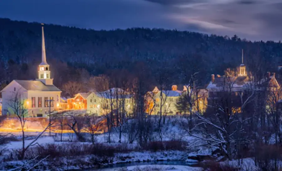 A snowy, serene village at dusk features a softly lit church with a tall steeple and charming, warm-lit houses against a backdrop of dark, wooded hills.