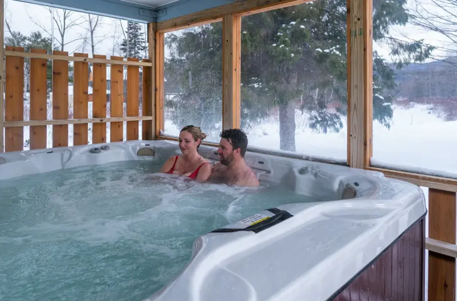 A couple relaxes in a hot tub on a snow-covered patio, surrounded by wooden railings. The scene is serene and cozy, contrasting warmth with a wintery backdrop.