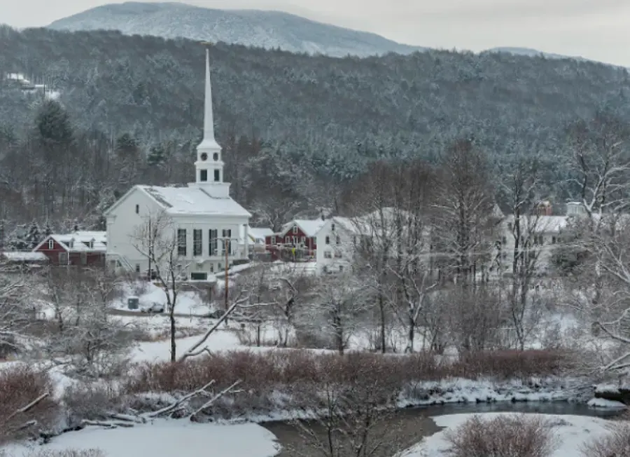 Snowy landscape with a white church and steeple amidst bare trees and houses, bordered by a winding river and hills in the background, evoking tranquility.