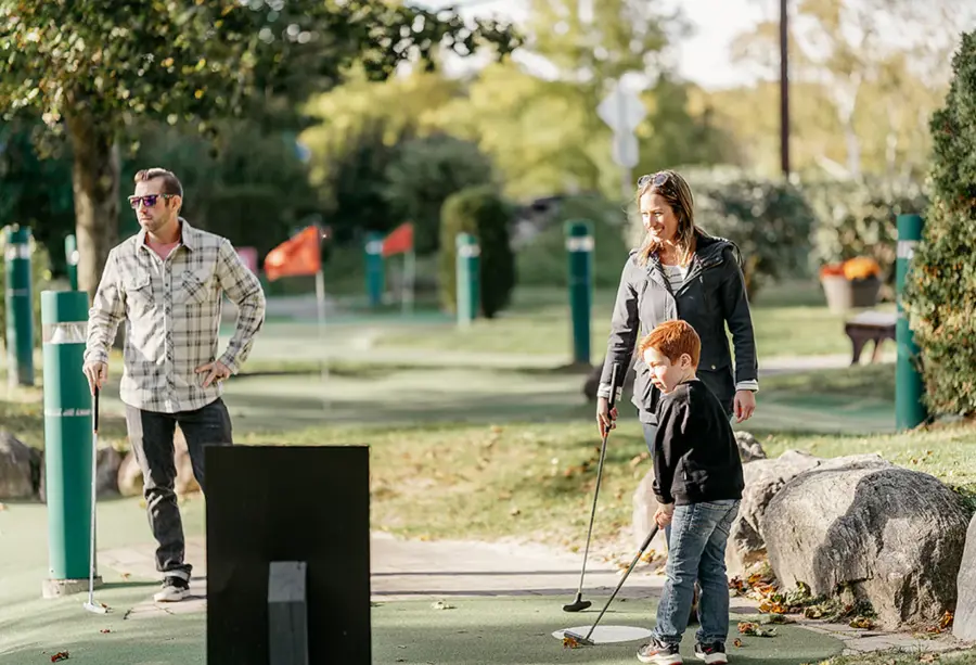 A family enjoys a sunny day at a mini-golf course. A man and a woman stand near a young boy holding a golf club, surrounded by trees and flags.