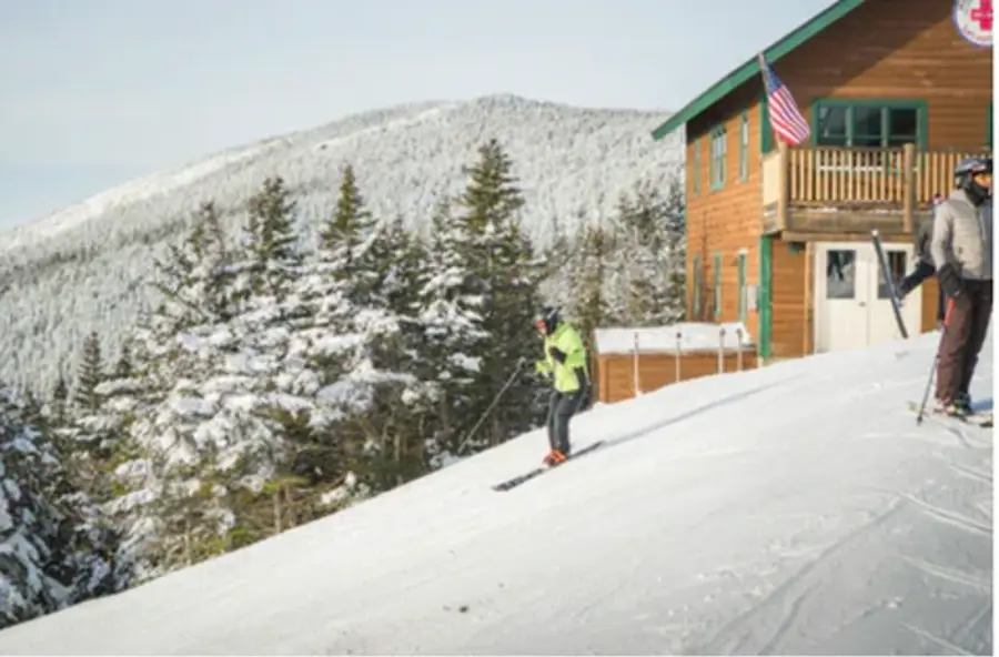 Two skiers pause on a snowy slope near a wooden cabin with an American flag, surrounded by snow-covered trees and mountains under a clear sky.