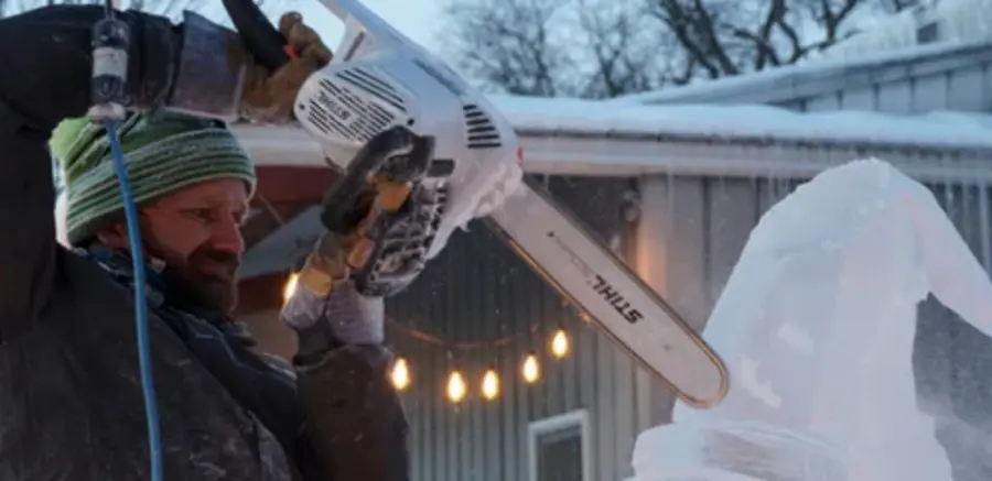 A man in a green beanie uses a chainsaw to carve an ice sculpture outdoors. String lights and a building are in the background, conveying a serene winter atmosphere.