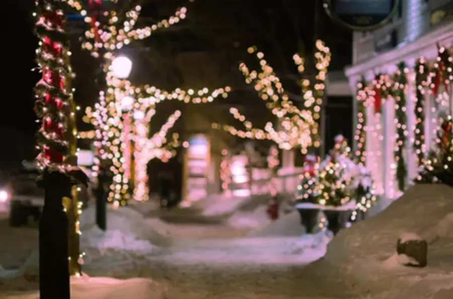 Snowy street adorned with festive string lights on trees and lamp posts. Decorated storefronts create a cozy, warm holiday ambiance at night.